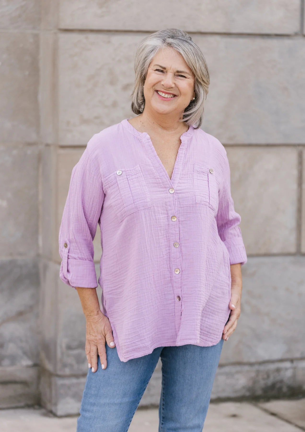 A person with long, blonde hair, wearing a ROXIE Woven Top by Shannon Passero made from quality materials stands against a plain background, showcasing a stylish look in their white button-up shirt with long sleeves.