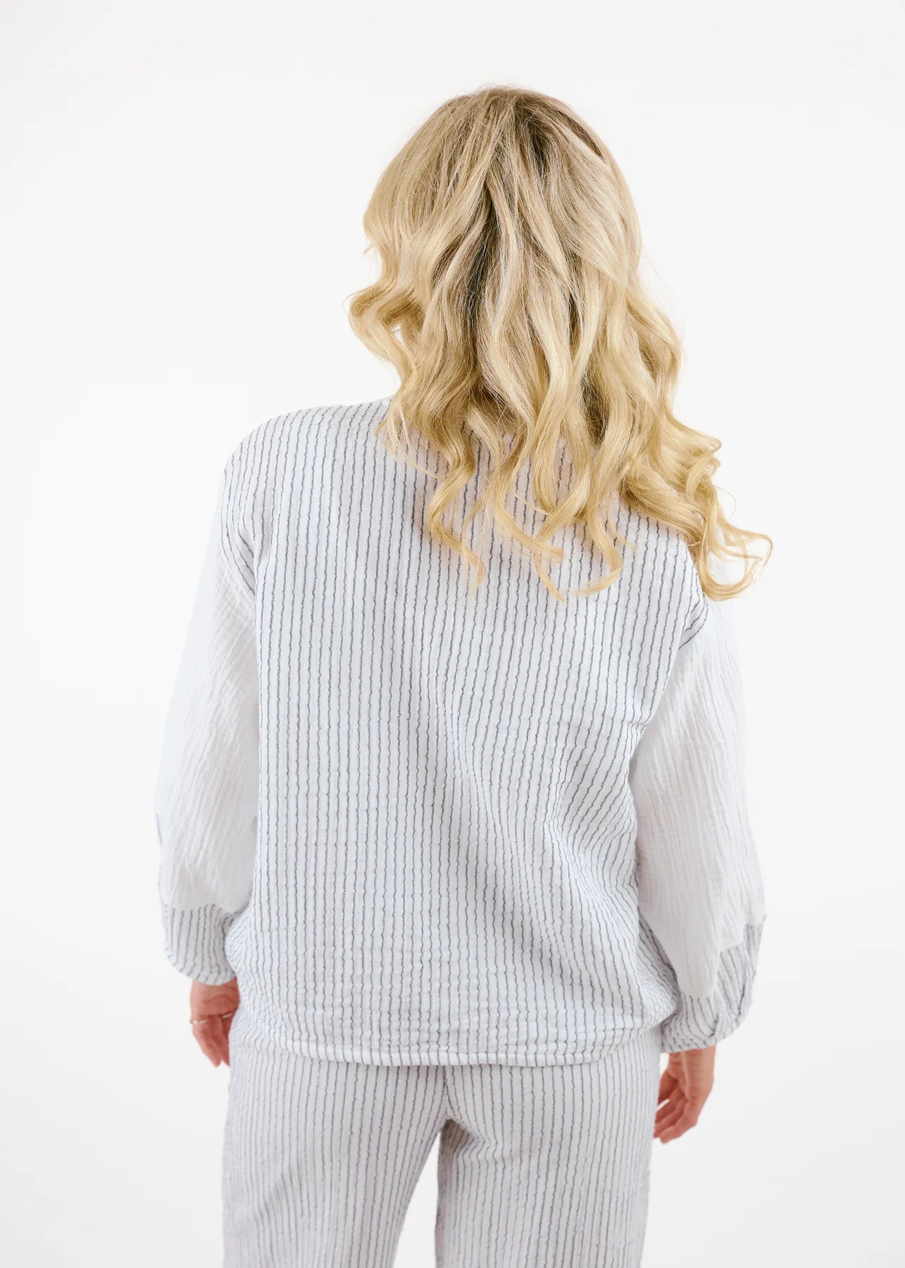 A person with long blonde hair is seen against a plain white background, wearing Shannon Passero's Kennedy Cardigan in white, featuring textured pin stripe detail and gray-striped pockets, along with matching pants.