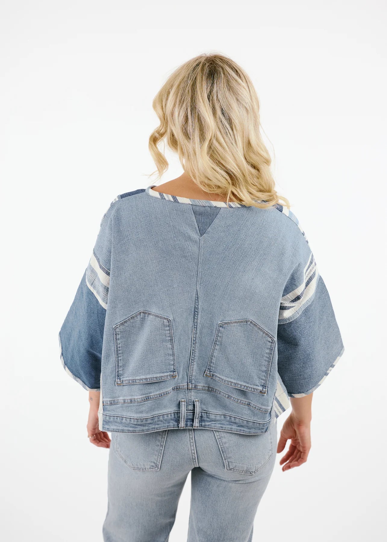 A woman wearing the Alex Top by Shannon Passero, featuring denim and stripes with light jeans crafted from upcycled denim, smiles while looking to the side against a stone wall.