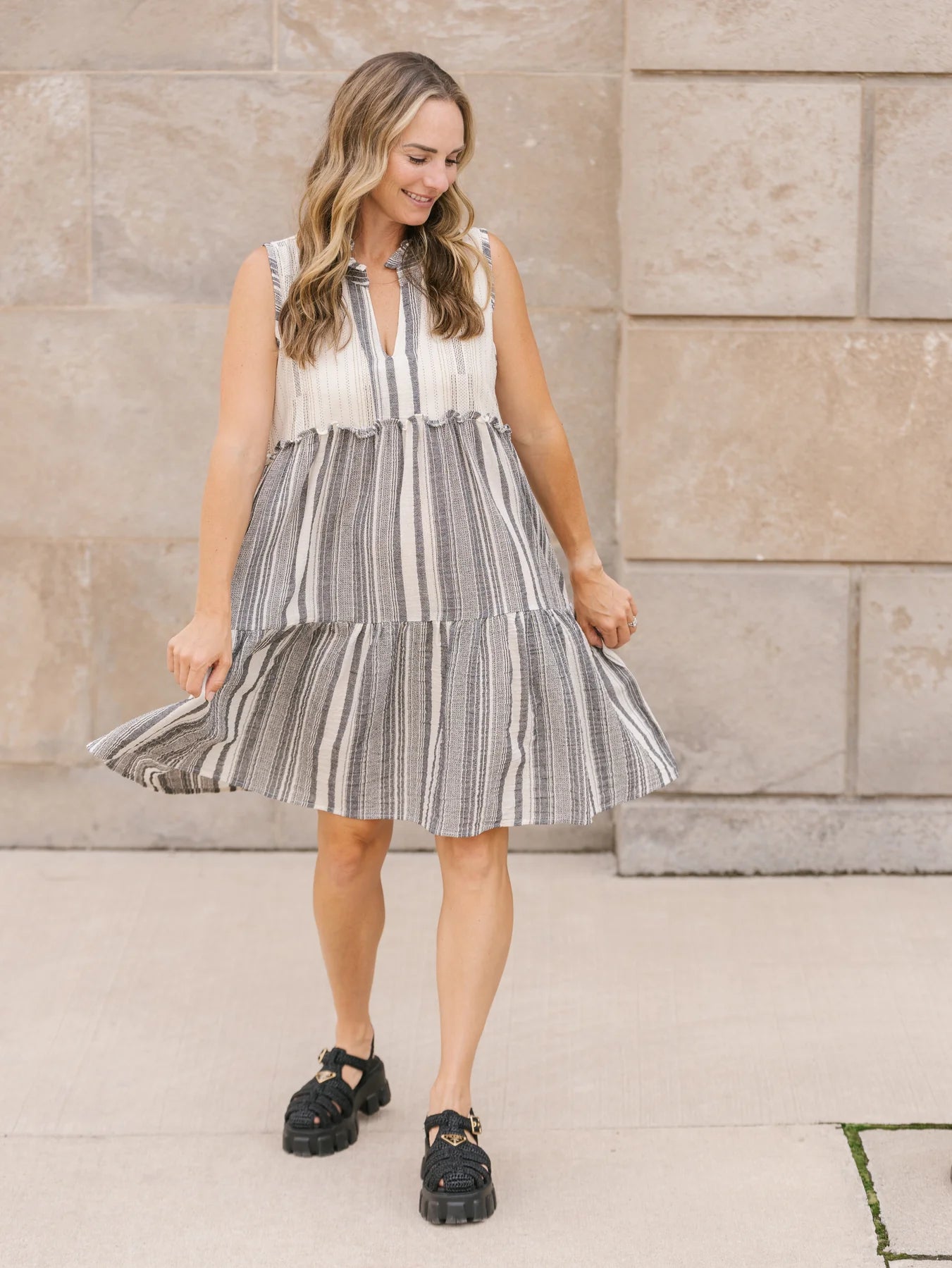 A person with long hair stands outdoors in a playful, v-neck, sleeveless Shannon Passero Whitby Tank Dress, smiling slightly.