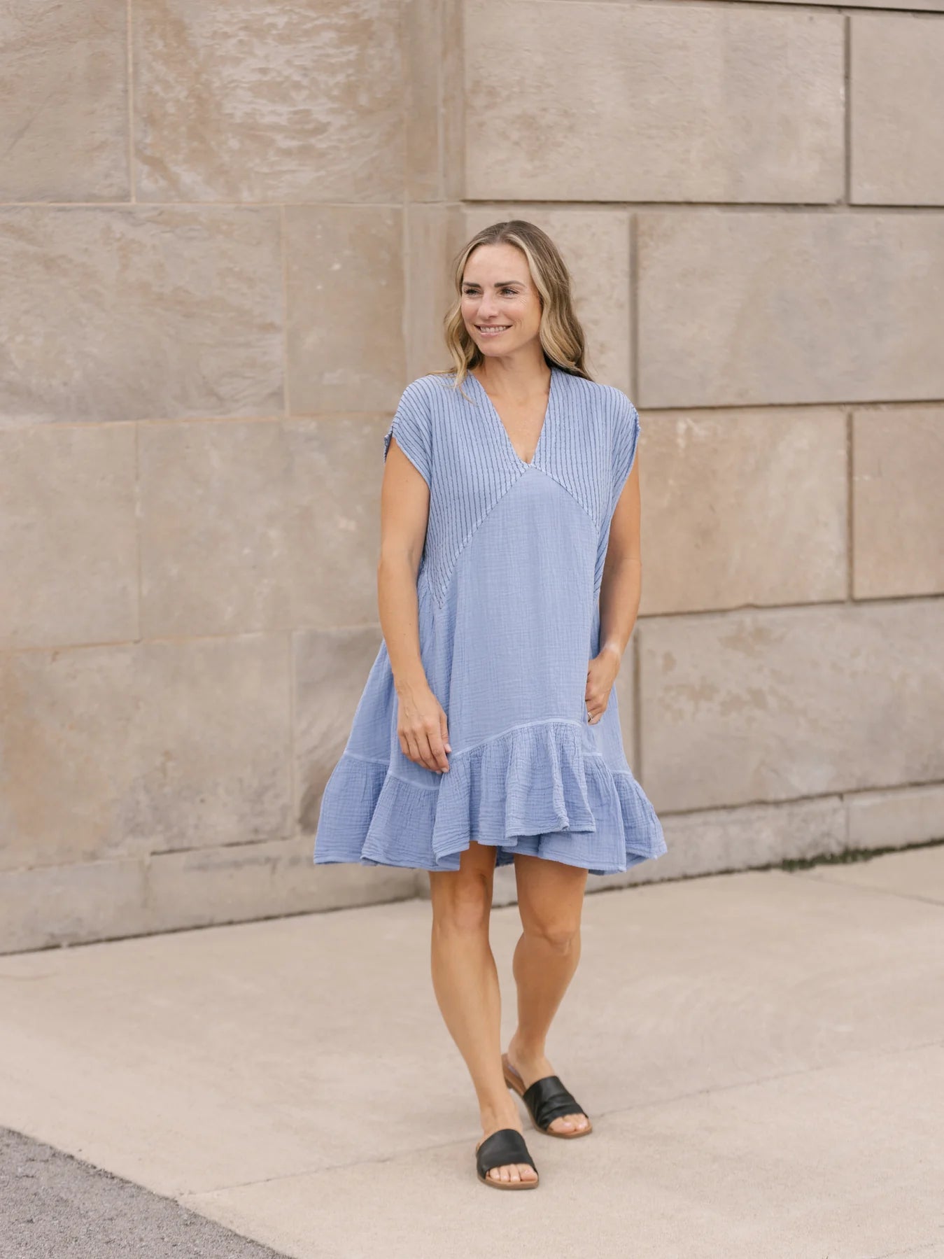 The woman in a flattering Shannon Passero Clover Dress and black sandals stands against a stone wall, showcasing the subtle ribbed detail that adds elegance to her ensemble.