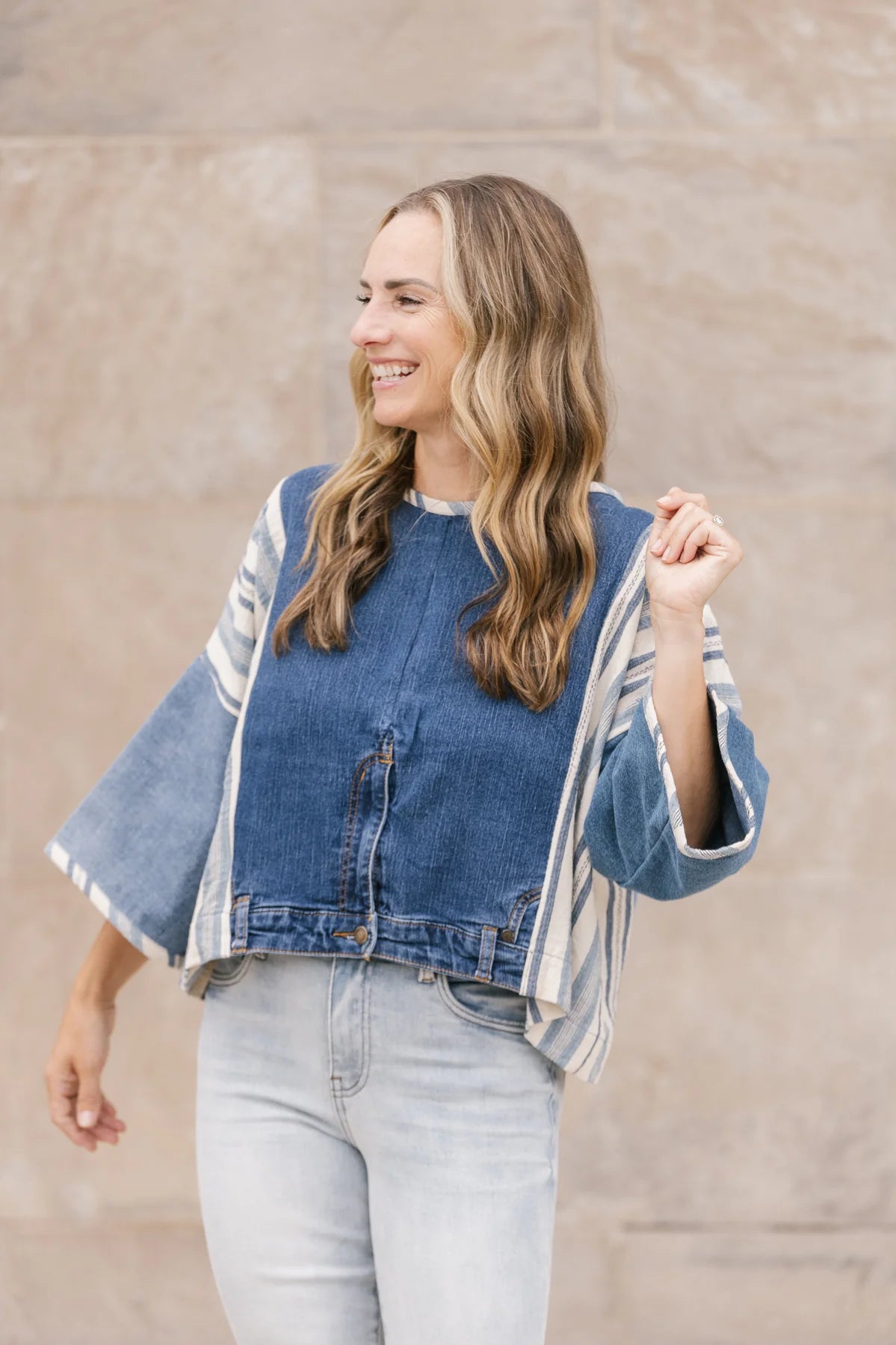 A woman wearing the Alex Top by Shannon Passero, featuring denim and stripes with light jeans crafted from upcycled denim, smiles while looking to the side against a stone wall.