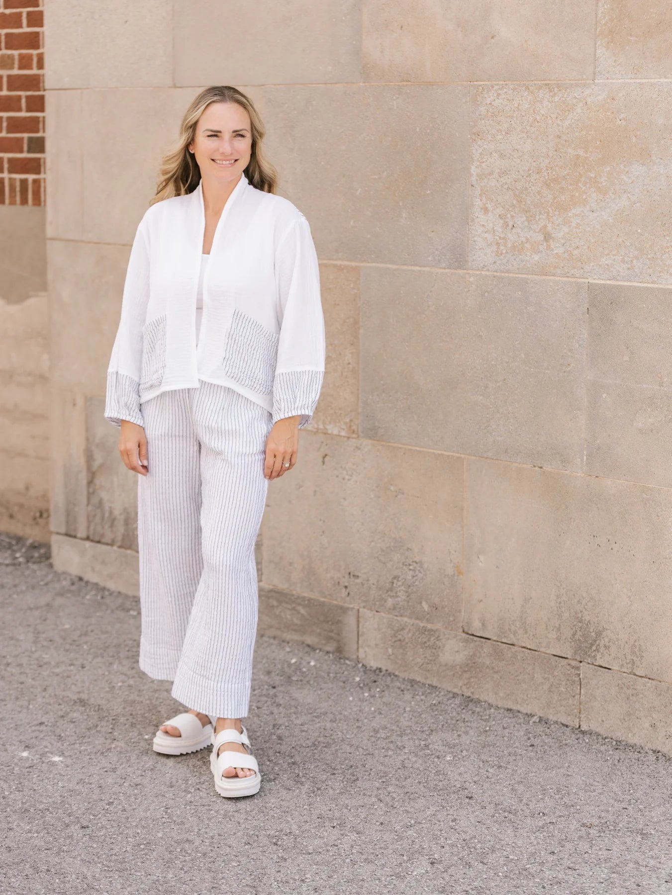 A person stands comfortably in Shannon Passero's Yucca Crop Pant, featuring light stripes, a loose fit, breathability, and front pockets, perfectly paired with a black top and flat sandals.