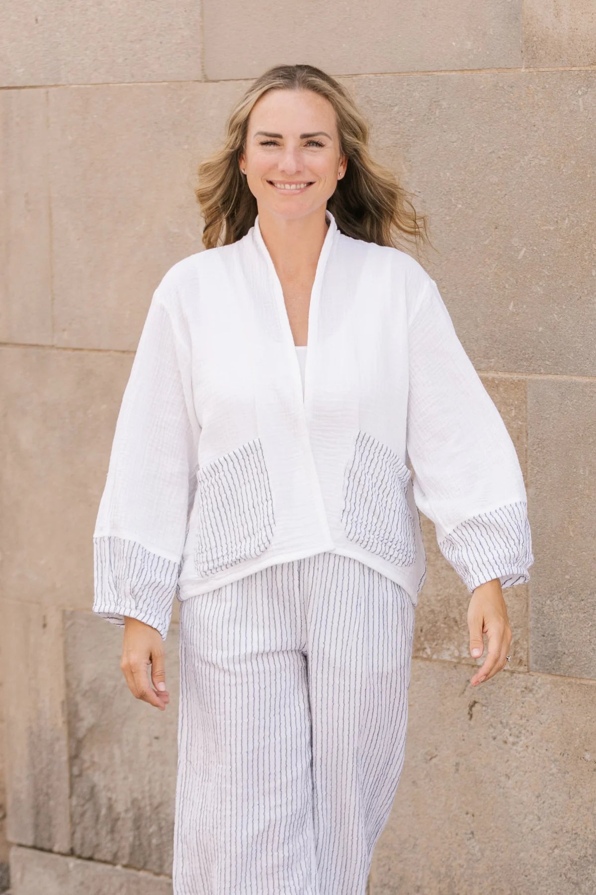 A person with long blonde hair is seen against a plain white background, wearing Shannon Passero's Kennedy Cardigan in white, featuring textured pin stripe detail and gray-striped pockets, along with matching pants.