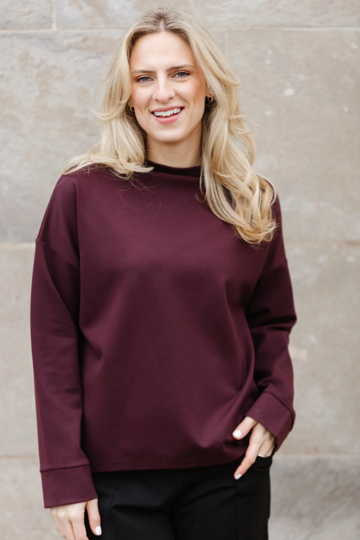 A person with long blonde hair, wearing a maroon Shannon Passero Wrenly Top crafted from high-quality materials, smiles at the camera in front of a textured stone wall.