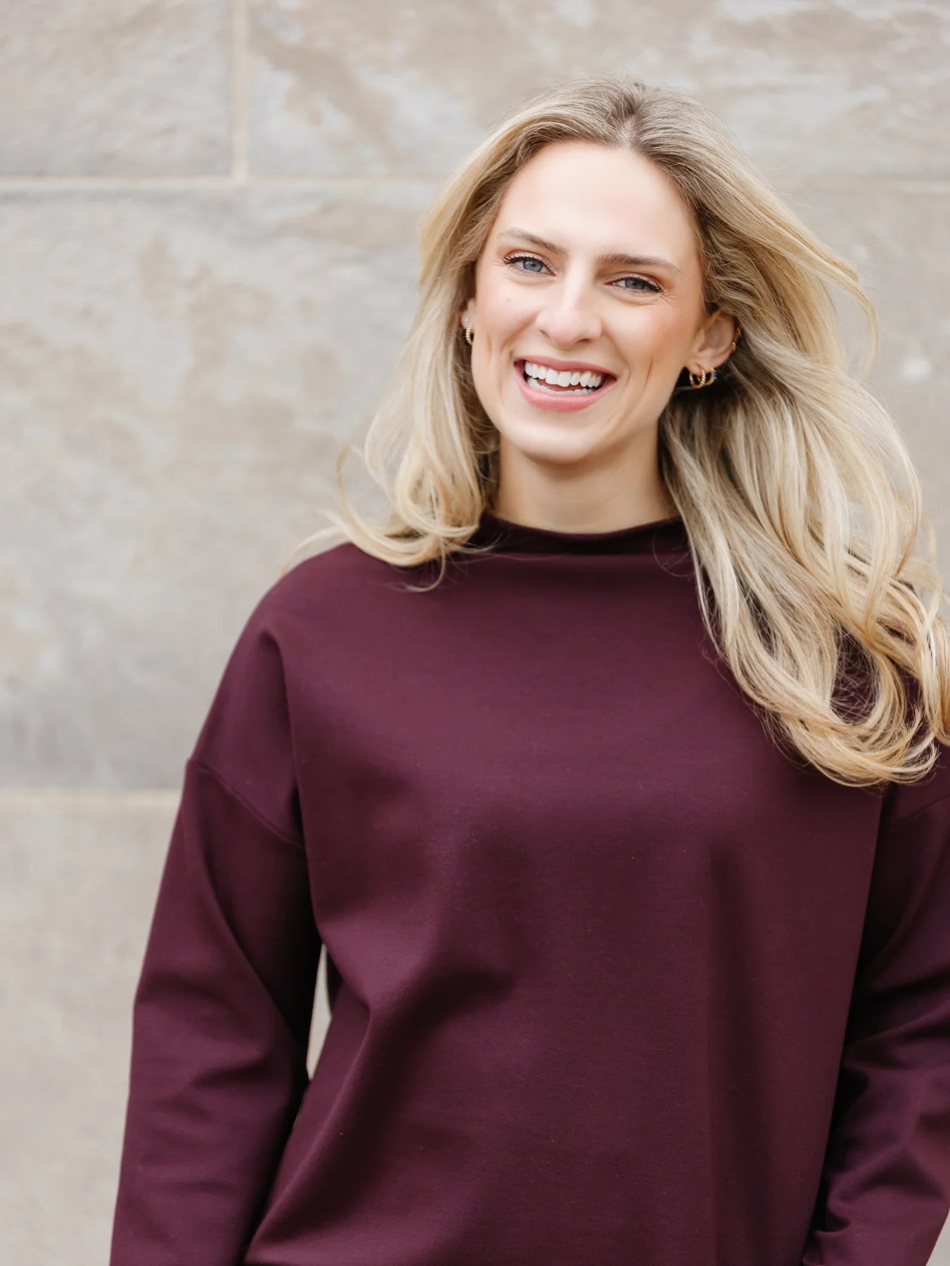 A woman with long blonde hair is wearing the Wrenly Top by Shannon Passero, a maroon sweater made of high-quality materials, paired with black pants. She is standing in front of a stone wall and smiling at the camera.