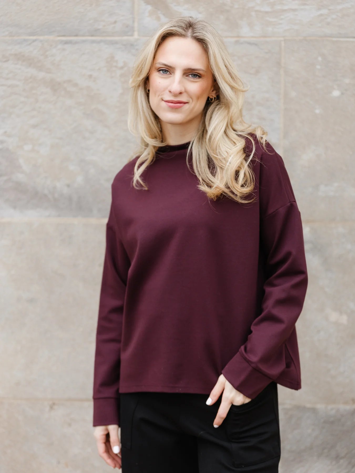 A woman with long blonde hair is wearing the Wrenly Top by Shannon Passero, a maroon sweater made of high-quality materials, paired with black pants. She is standing in front of a stone wall and smiling at the camera.