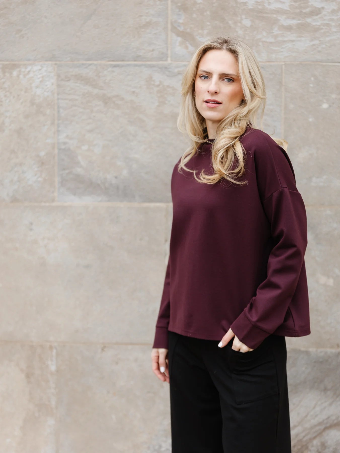 A woman with long blonde hair is wearing the Wrenly Top by Shannon Passero, a maroon sweater made of high-quality materials, paired with black pants. She is standing in front of a stone wall and smiling at the camera.