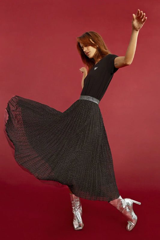 A person poses against a red backdrop, wearing a black top and the Pleated Plumetis Skirt by Molly Bracken, adorned with polka dot feathers, completed by silver heeled boots.