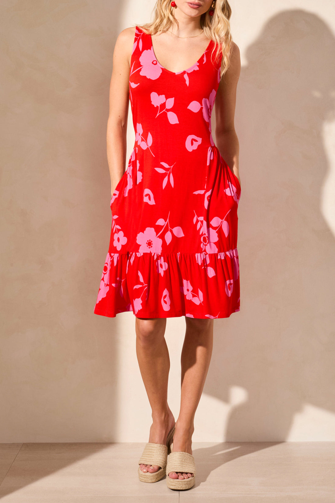 A woman poses against a light wall, highlighting a stylish Tribal Printed Sleeveless Dress featuring blue with white floral patterns, expertly paired with white wedge sandals.