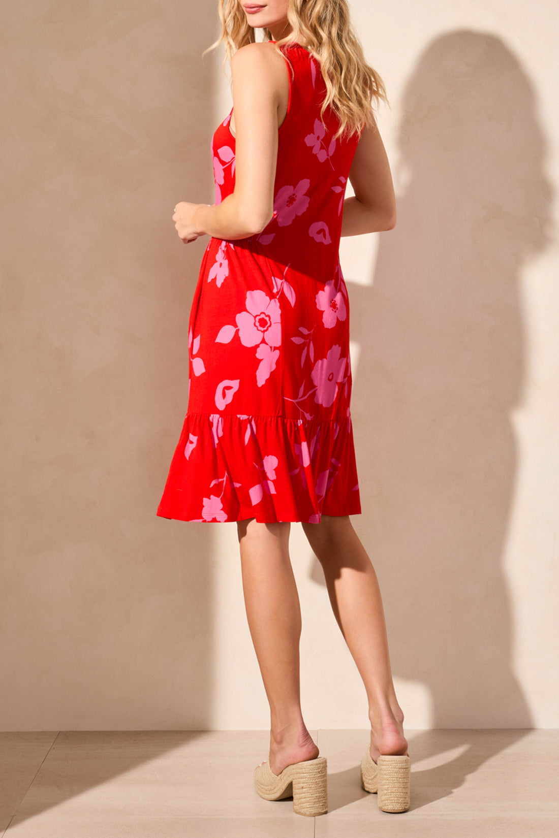 A woman poses against a light wall, highlighting a stylish Tribal Printed Sleeveless Dress featuring blue with white floral patterns, expertly paired with white wedge sandals.