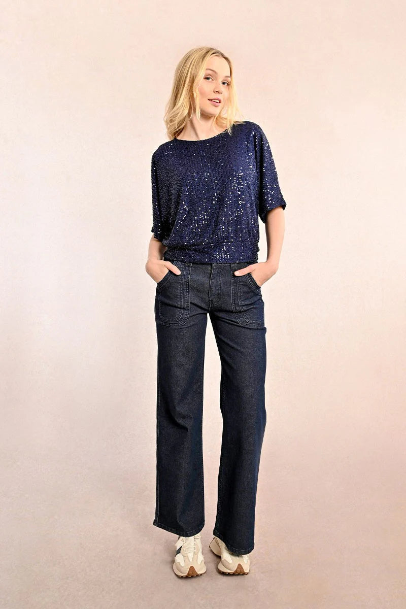 A woman in a chic Sequin Open Back Top by Molly Bracken, paired with jeans, poses with her hands in her pockets against a neutral background.