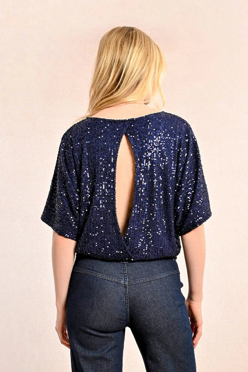 A woman in a chic Sequin Open Back Top by Molly Bracken, paired with jeans, poses with her hands in her pockets against a neutral background.