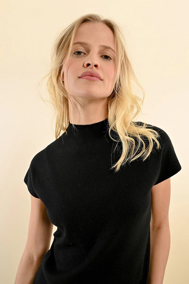 A person with long blonde hair is standing with their back to the camera, wearing a Short Sleeve Ribbed Sweater from Molly Bracken and blue jeans against a neutral background.