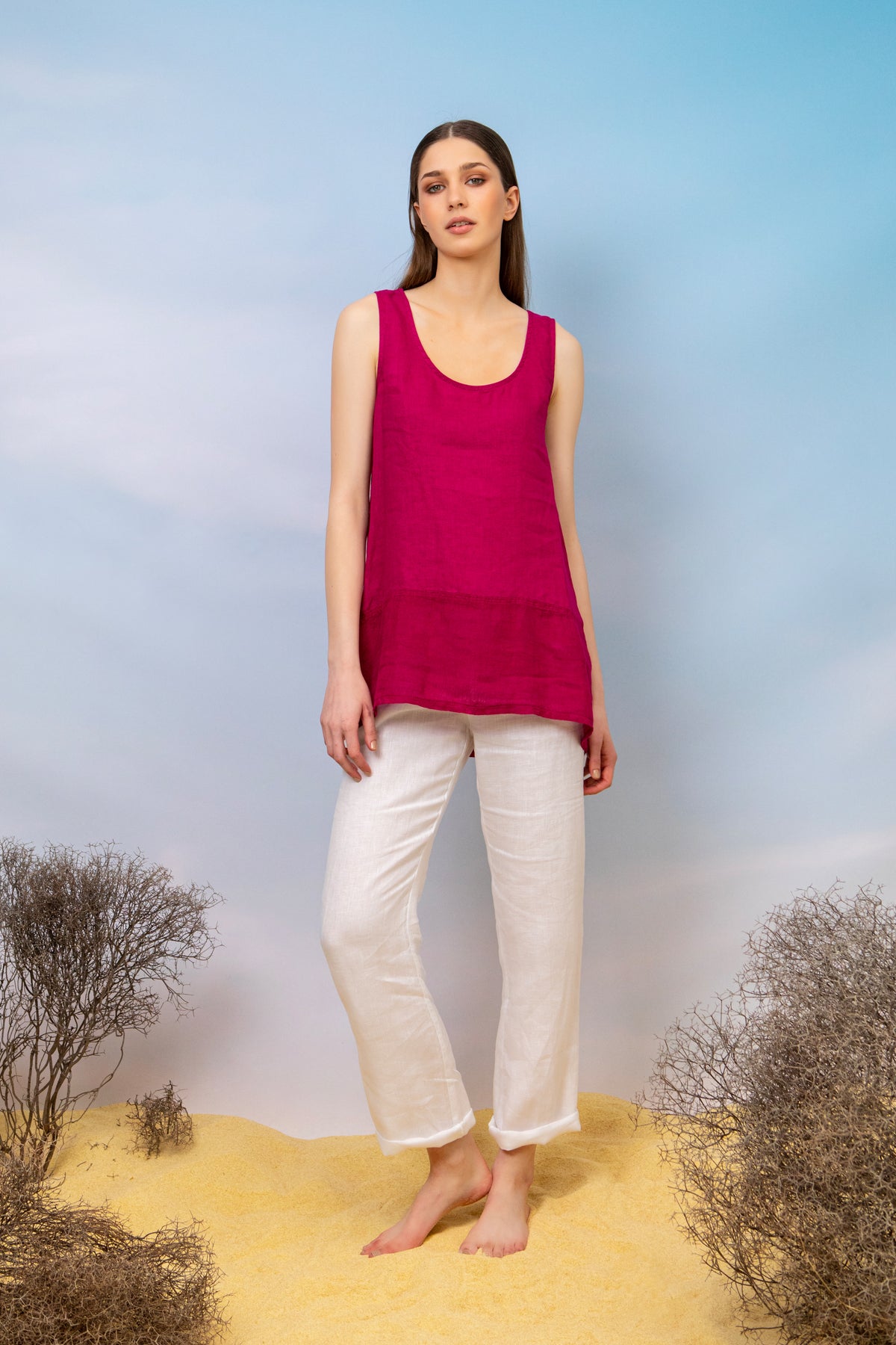 A woman stands barefoot on light yellow sand against a gradient blue sky, wearing a DS Collection Sleeveless Linen Blouse in magenta and white pants, with sparse bushes in the foreground. Perfect for any summer wardrobe, this breathable top offers both comfort and style amidst the serene landscape.