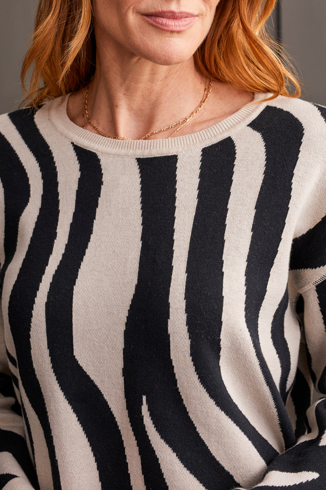 A person wearing a Tribal Long Sleeve Crew Neck Reversible Sweater with black floral patterns, matched with brown pants and black ankle boots, stands in front of a paneled gray wall.