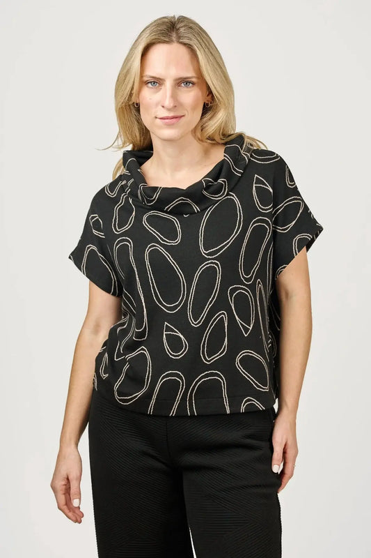 A woman with blonde hair is wearing a fashion-forward Harlee Top by Shannon Passero, featuring an abstract oval pattern, paired with black pants, and standing against a plain background.