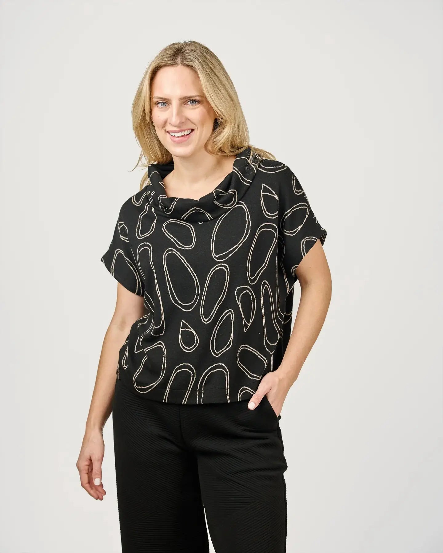 A woman with blonde hair is wearing a fashion-forward Harlee Top by Shannon Passero, featuring an abstract oval pattern, paired with black pants, and standing against a plain background.