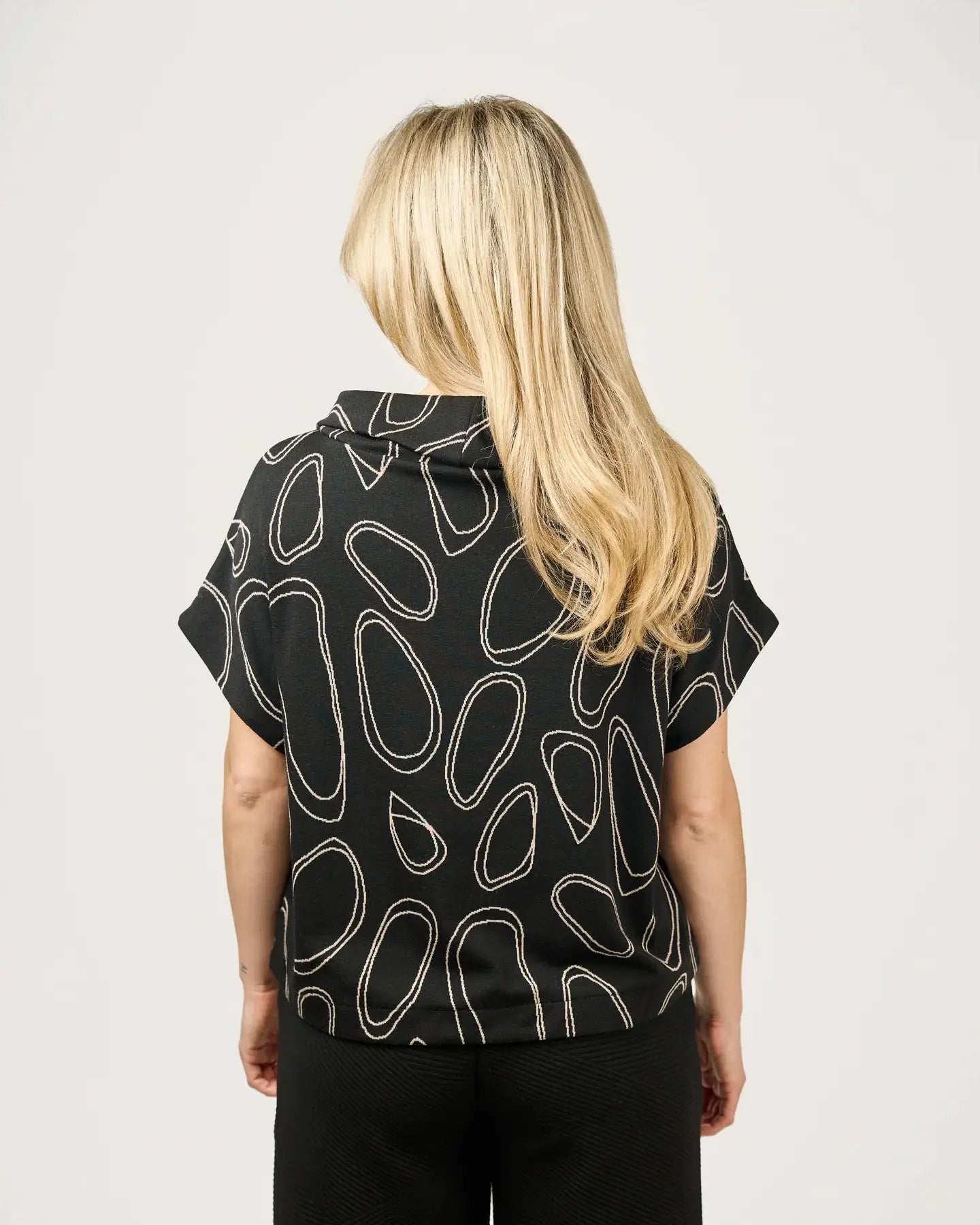 A woman with blonde hair is wearing a fashion-forward Harlee Top by Shannon Passero, featuring an abstract oval pattern, paired with black pants, and standing against a plain background.