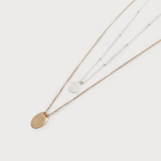 The Caracol 2 Layer Chains with Small Charms feature two delicate chains adorned with small circular pendants and flat metallic charms, available in both gold and silver finishes. The elegant necklaces are displayed on a plain white background.