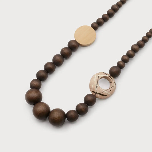 The Caracol Wooden Beads Necklace showcases dark brown wooden beads, accented by a large circular light wood bead with etched designs, and includes a flat, round gold bead.