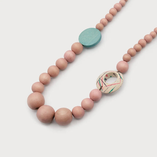 The Wooden Beads Necklace by Caracol showcases round pink beads, a single light blue bead, and an elliptical white bead with multicolored streaks, all accented with painted wood pieces.