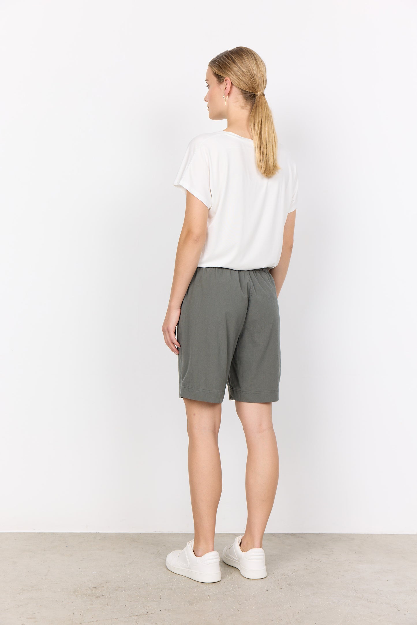 A person stands sideways in a cozy white cotton t-shirt, paired with stylish Cissie 2 Shorts from Soya Concepts and crisp white sneakers, all set against a plain white background.