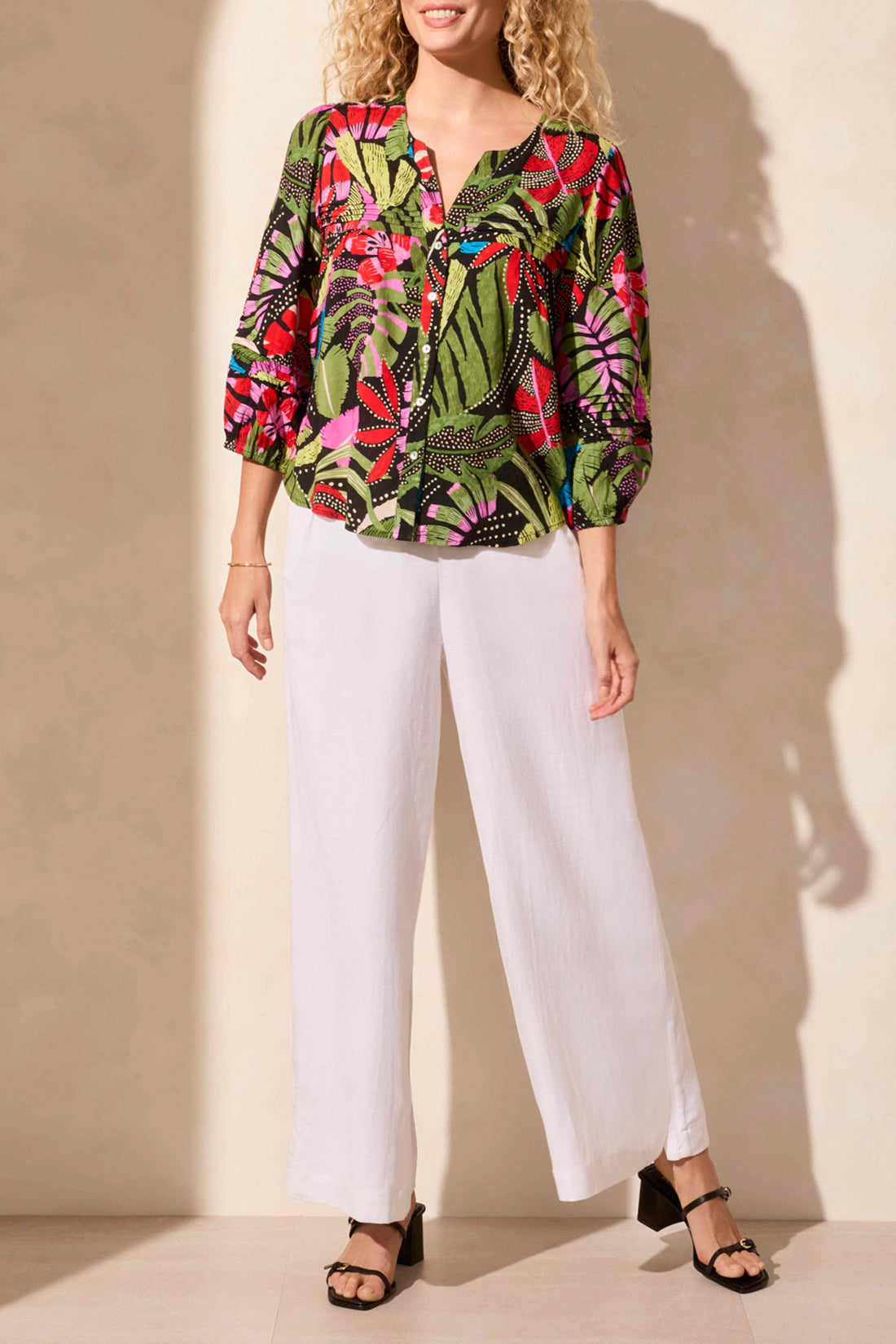 A woman stands against a light backdrop wearing Tribal's colorful 3/4 Sleeve Button Front Blouse with pleats, featuring abstract leaf designs, paired with white pants.