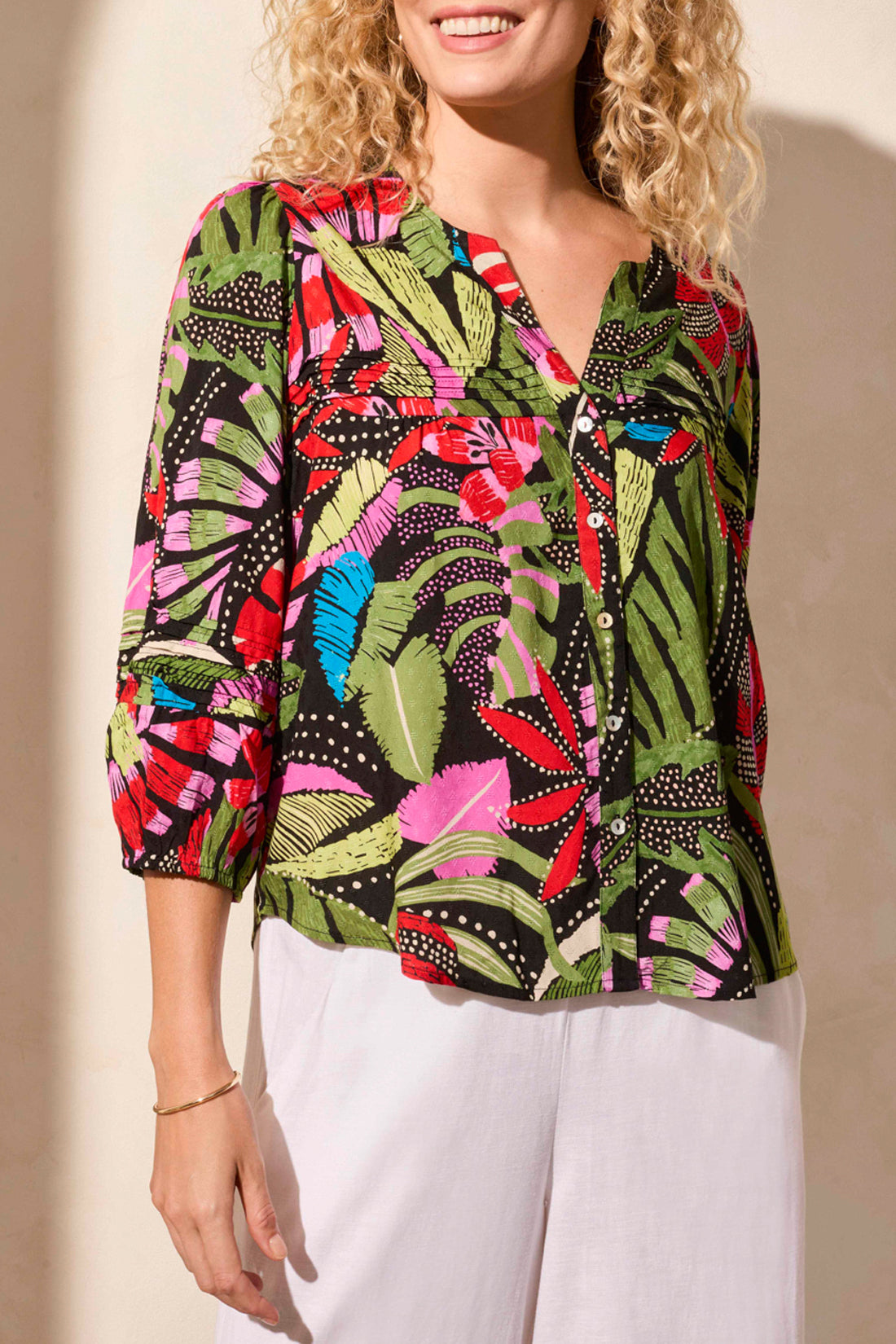 A woman stands against a light backdrop wearing Tribal's colorful 3/4 Sleeve Button Front Blouse with pleats, featuring abstract leaf designs, paired with white pants.