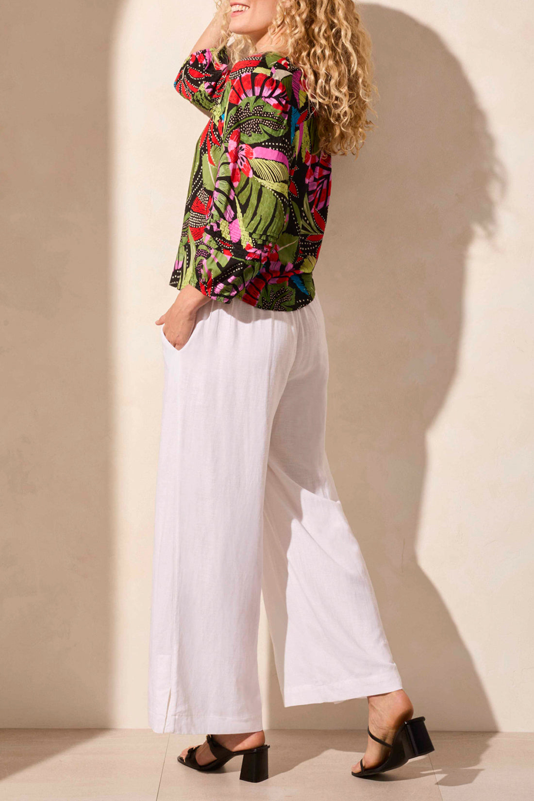 A woman stands against a light backdrop wearing Tribal's colorful 3/4 Sleeve Button Front Blouse with pleats, featuring abstract leaf designs, paired with white pants.