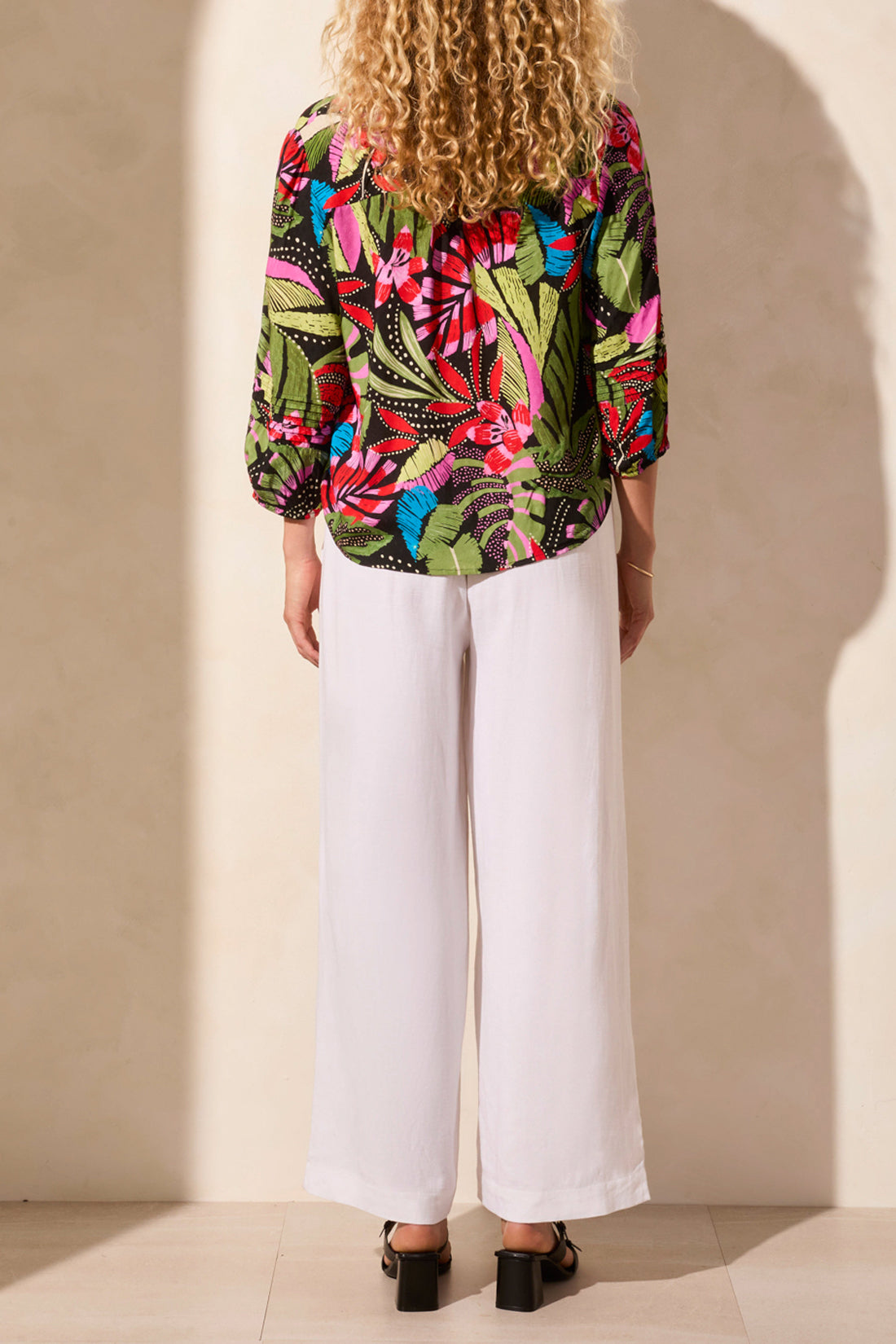 A woman stands against a light backdrop wearing Tribal's colorful 3/4 Sleeve Button Front Blouse with pleats, featuring abstract leaf designs, paired with white pants.