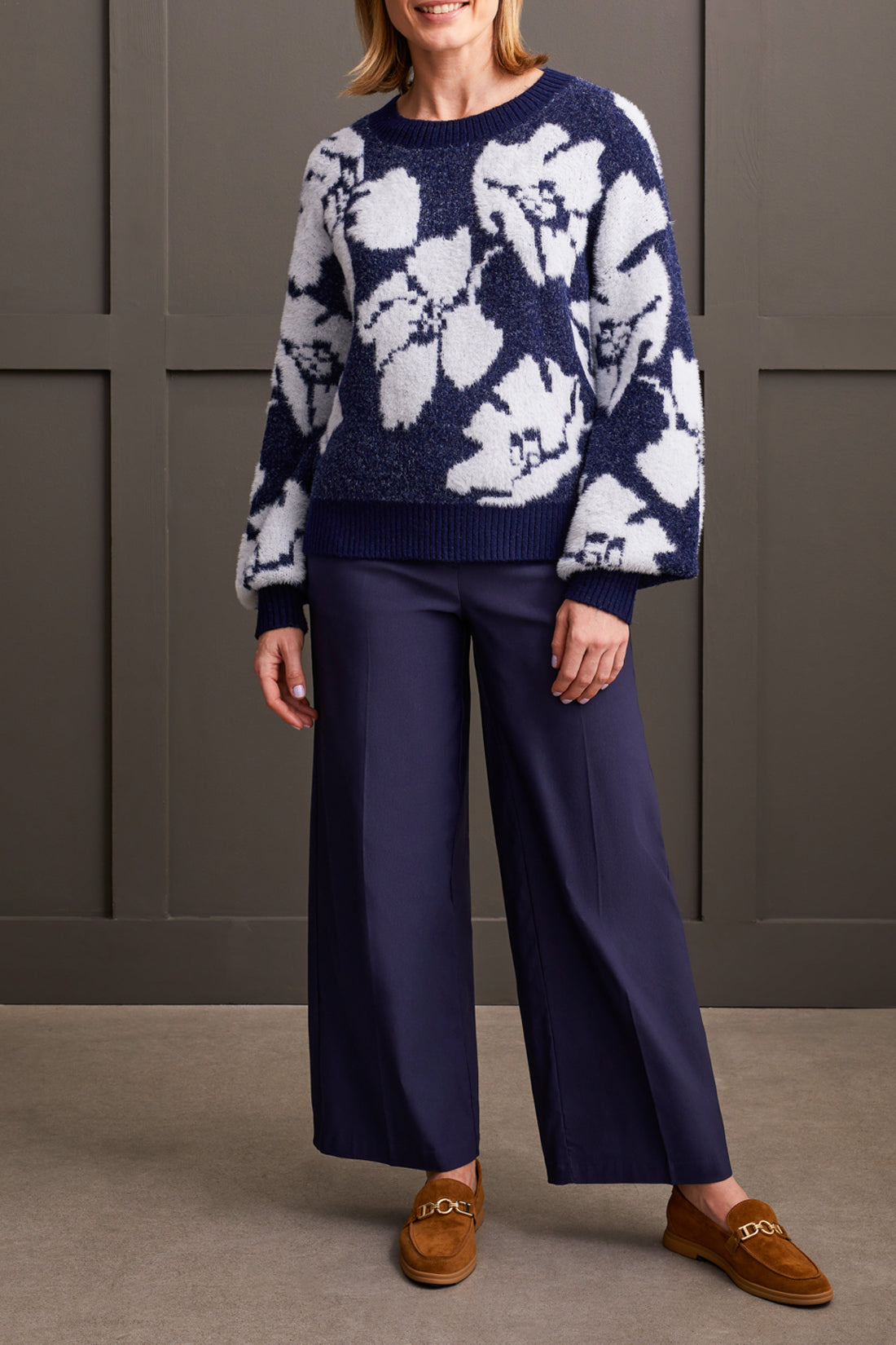 A person clad in the comfortable Oversized Floral Crew Neck Sweater by Tribal, coupled with wide-leg navy pants and brown loafers, leans effortlessly against a paneled wall.