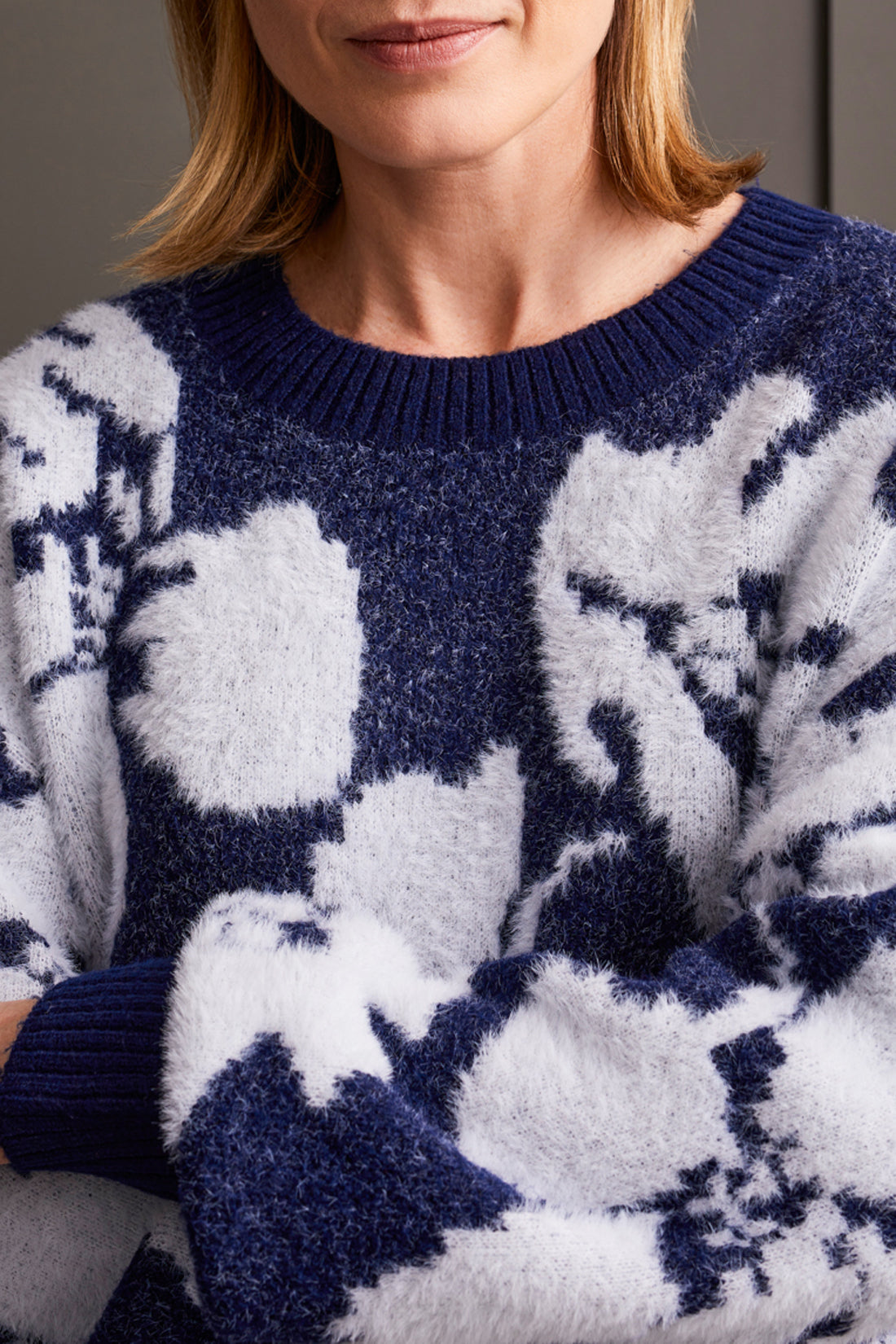 A person clad in the comfortable Oversized Floral Crew Neck Sweater by Tribal, coupled with wide-leg navy pants and brown loafers, leans effortlessly against a paneled wall.