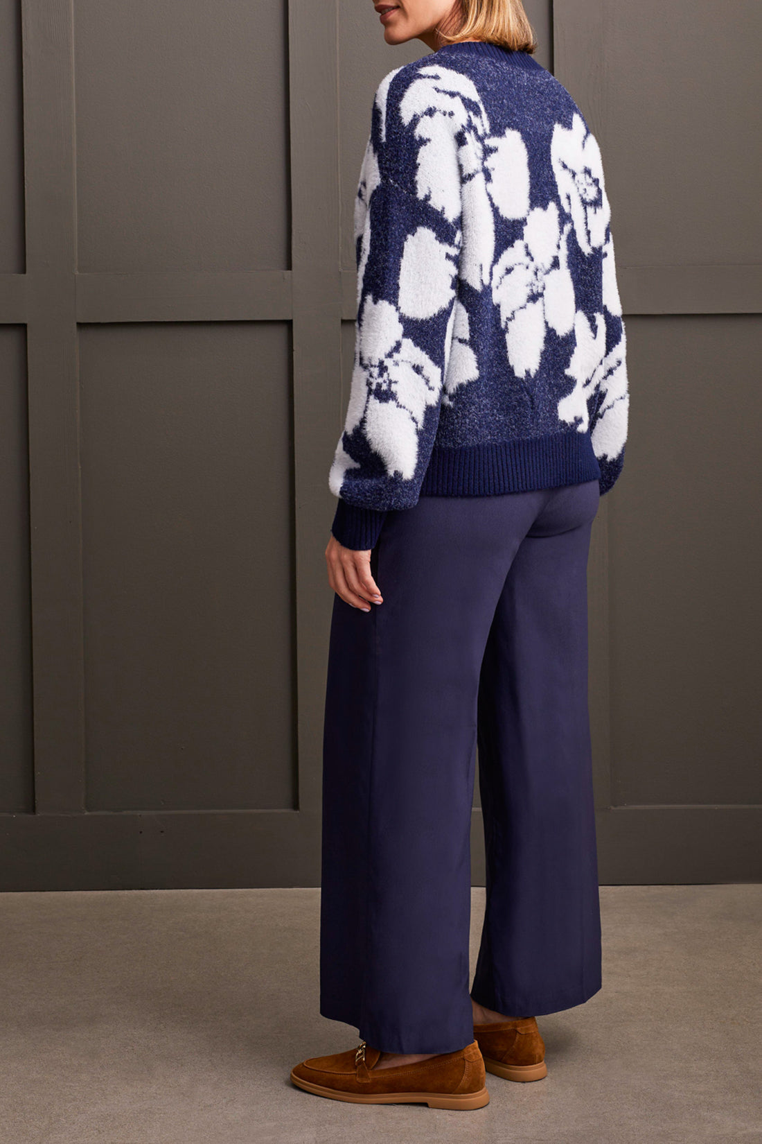 A person clad in the comfortable Oversized Floral Crew Neck Sweater by Tribal, coupled with wide-leg navy pants and brown loafers, leans effortlessly against a paneled wall.