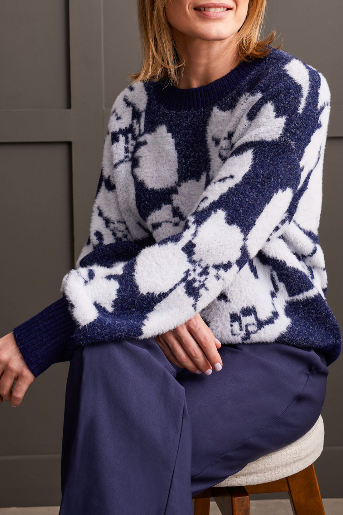 A person clad in the comfortable Oversized Floral Crew Neck Sweater by Tribal, coupled with wide-leg navy pants and brown loafers, leans effortlessly against a paneled wall.