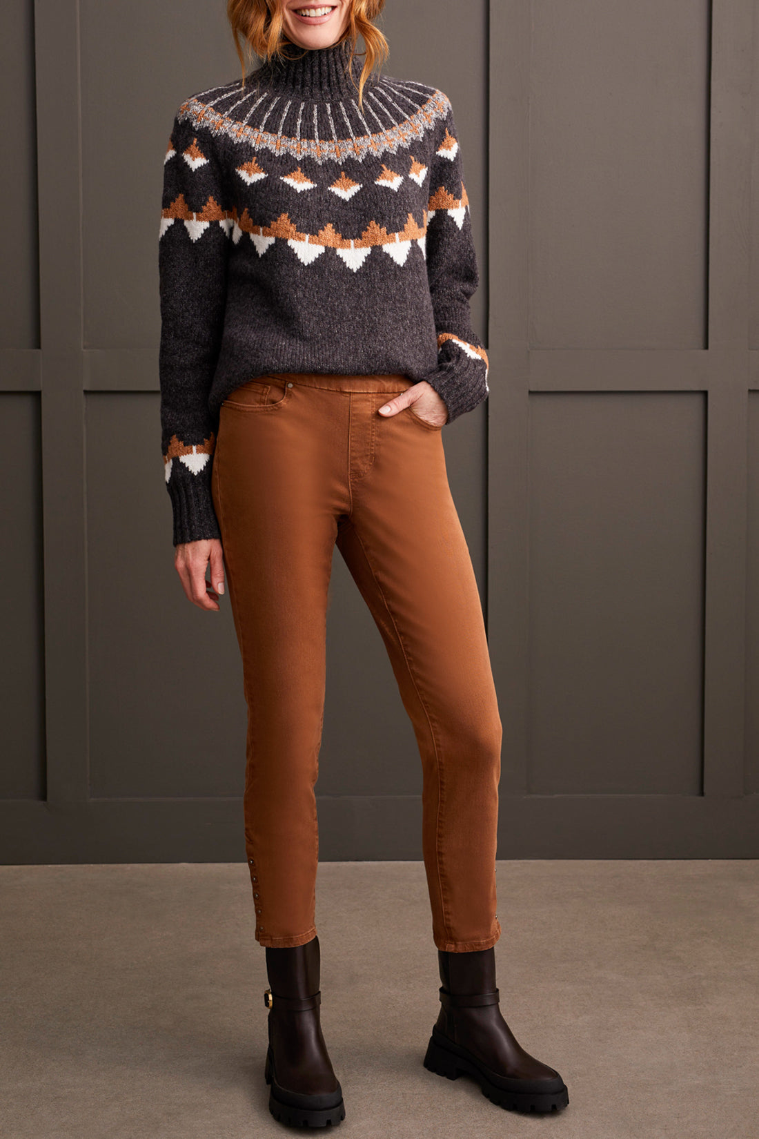 A person wearing a patterned sweater, Tribal's Pull on Ankle Pant with Hem Vent, and dark boots stands against a paneled wall.