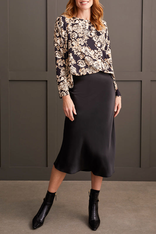 A person wearing the Tribal Crew Neck Top with Curved Hem, featuring a stylish patterned design, paired with a black knee-length skirt and black ankle boots, stands against a dark paneled wall—perfect for any fall wardrobe.