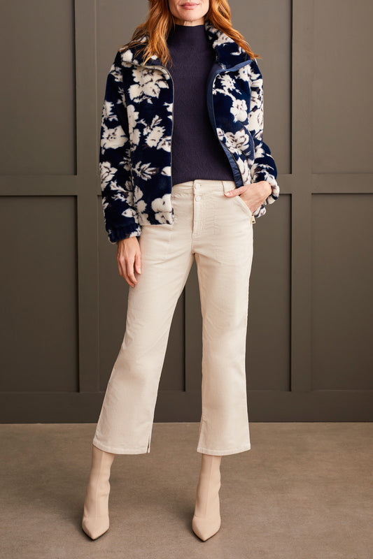 A person wearing the Tribal Zip up Bomber with Lining featuring a floral pattern, along with a dark turtleneck, light-colored pants, and beige boots, stands against a paneled wall.