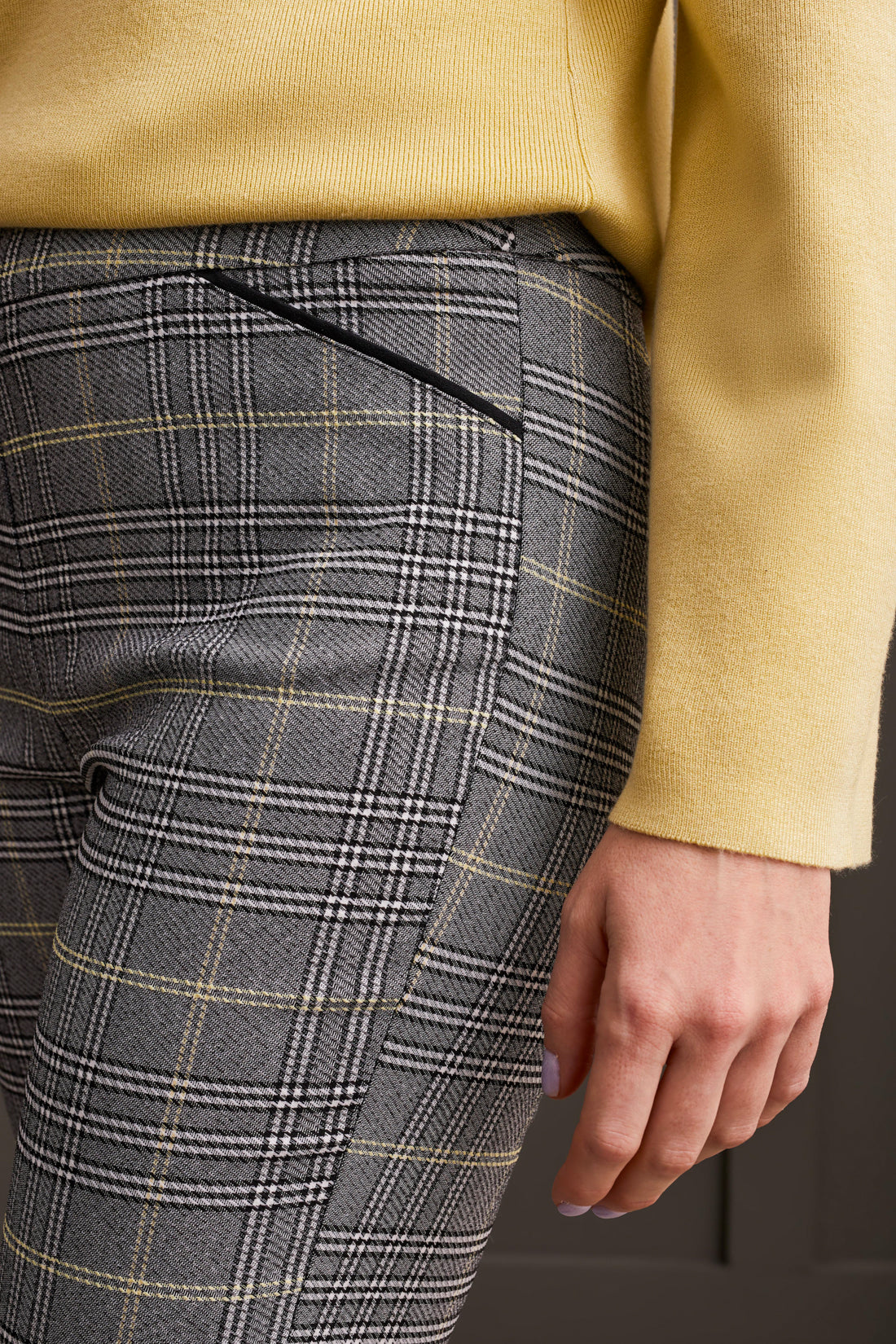 A person wearing a yellow long-sleeve top, comfortable gray plaid Pull on Ankle Pant by Tribal, and black ankle boots stands against a dark-paneled wall.