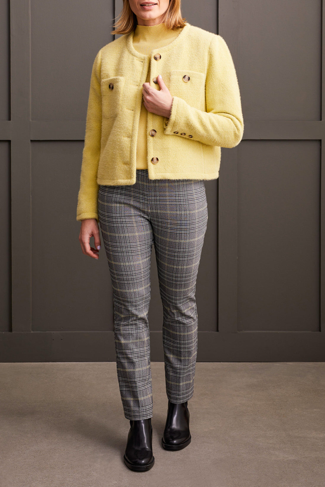 A person wearing a yellow long-sleeve top, comfortable gray plaid Pull on Ankle Pant by Tribal, and black ankle boots stands against a dark-paneled wall.