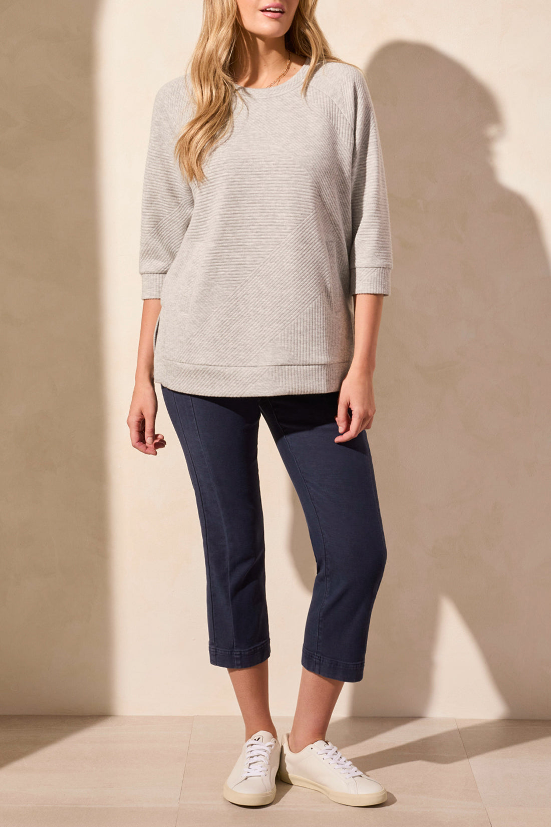 A person is wearing a light gray 3/4 Sleeve Tunic with Pockets by Tribal, paired with dark blue capri pants and white sneakers, standing against a neutral background.