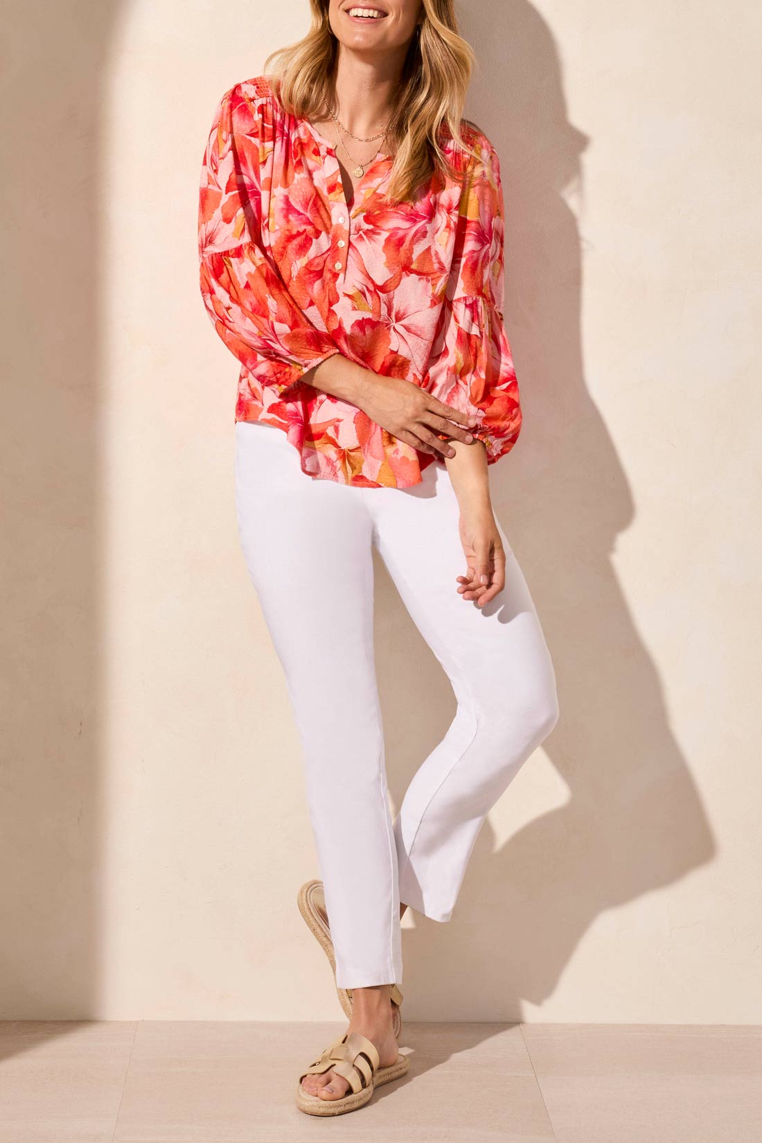 Someone in the Tribal 3/4 Sleeve Blouse with Smocked Yoke, featuring a vibrant floral pattern in pink and orange, paired with white pants against a neutral background.