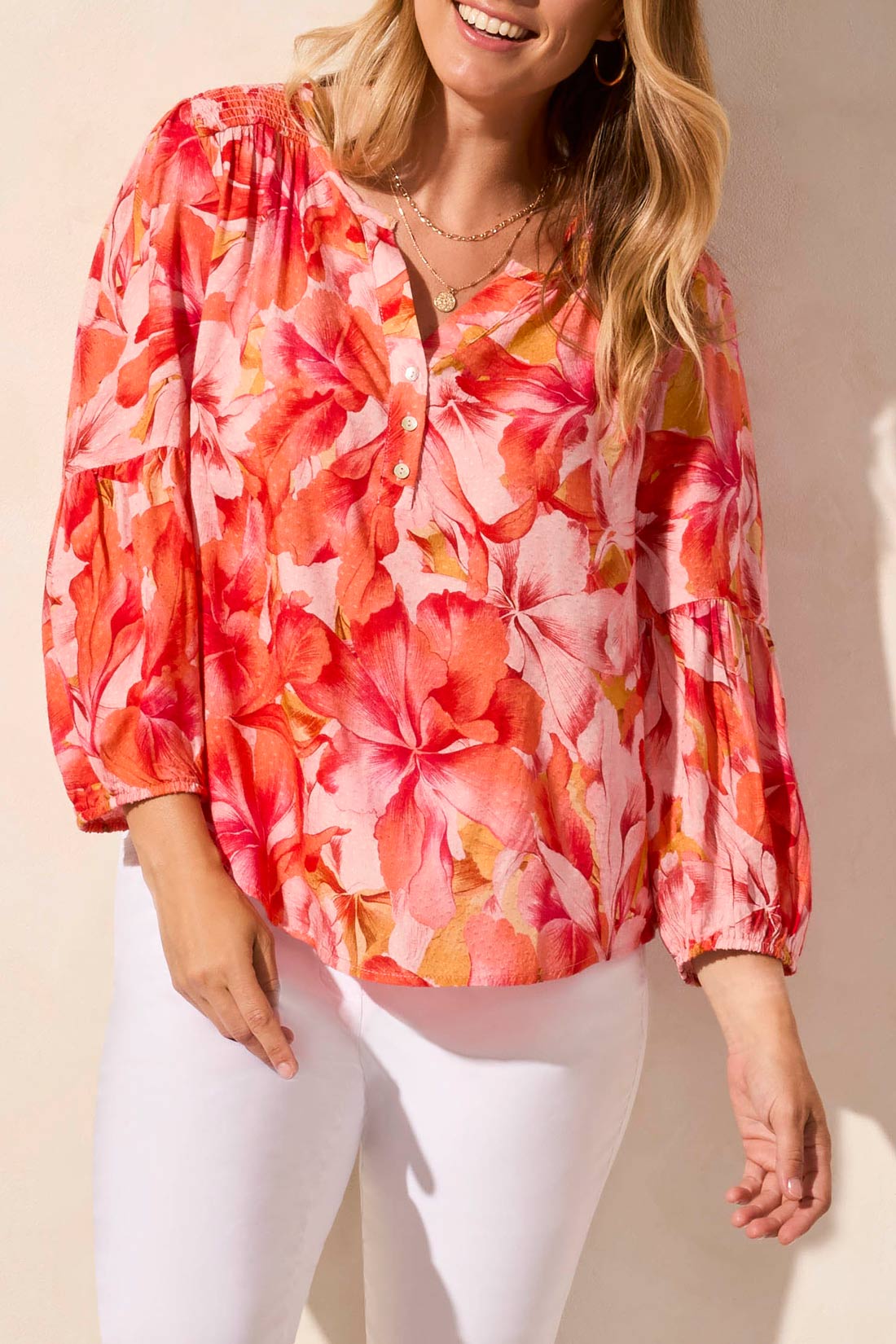 Someone in the Tribal 3/4 Sleeve Blouse with Smocked Yoke, featuring a vibrant floral pattern in pink and orange, paired with white pants against a neutral background.