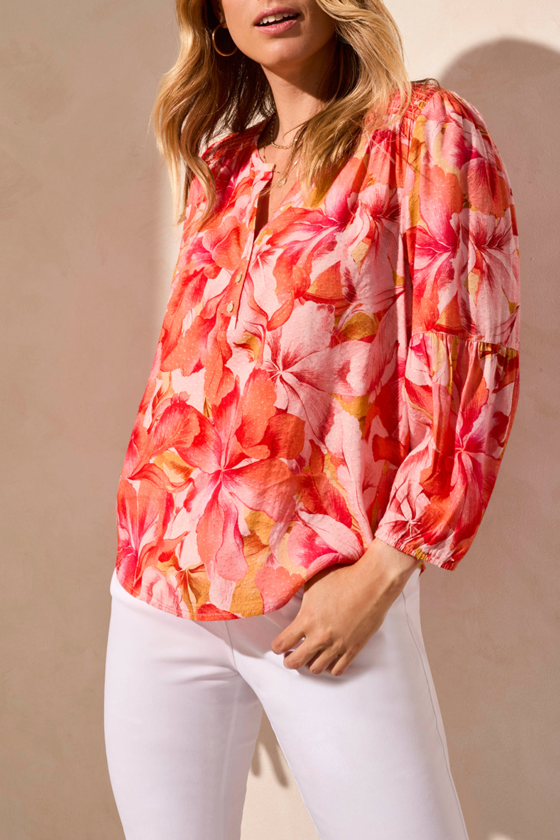 Someone in the Tribal 3/4 Sleeve Blouse with Smocked Yoke, featuring a vibrant floral pattern in pink and orange, paired with white pants against a neutral background.