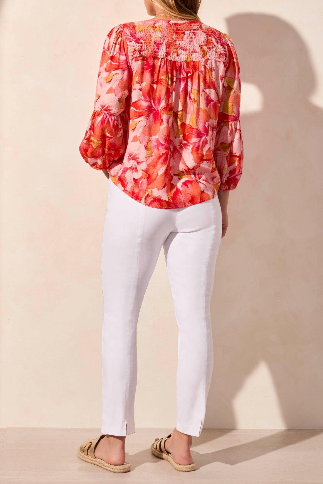 Someone in the Tribal 3/4 Sleeve Blouse with Smocked Yoke, featuring a vibrant floral pattern in pink and orange, paired with white pants against a neutral background.