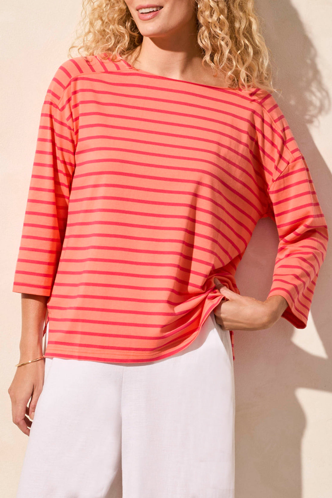 A person wearing a stylish Tribal 3/4 Sleeve Boat Neck Top in coral and pink stripes paired with white pants stands against a beige background.