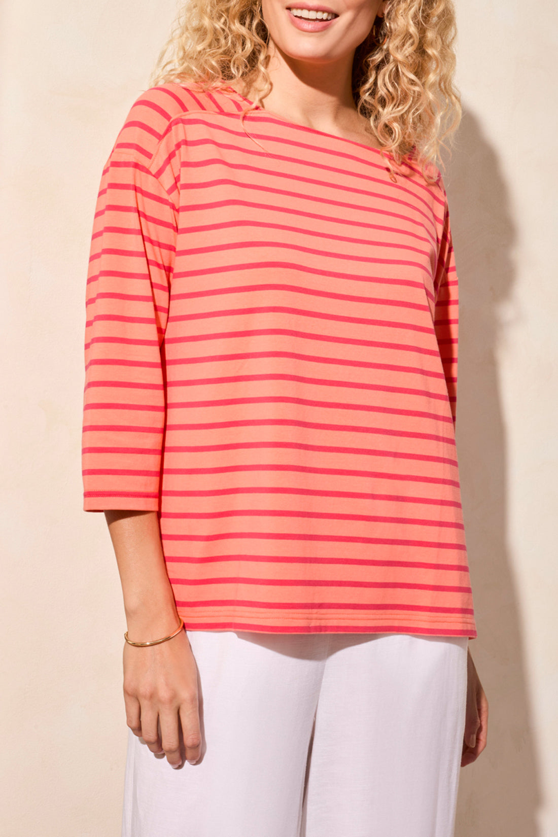 A person wearing a stylish Tribal 3/4 Sleeve Boat Neck Top in coral and pink stripes paired with white pants stands against a beige background.