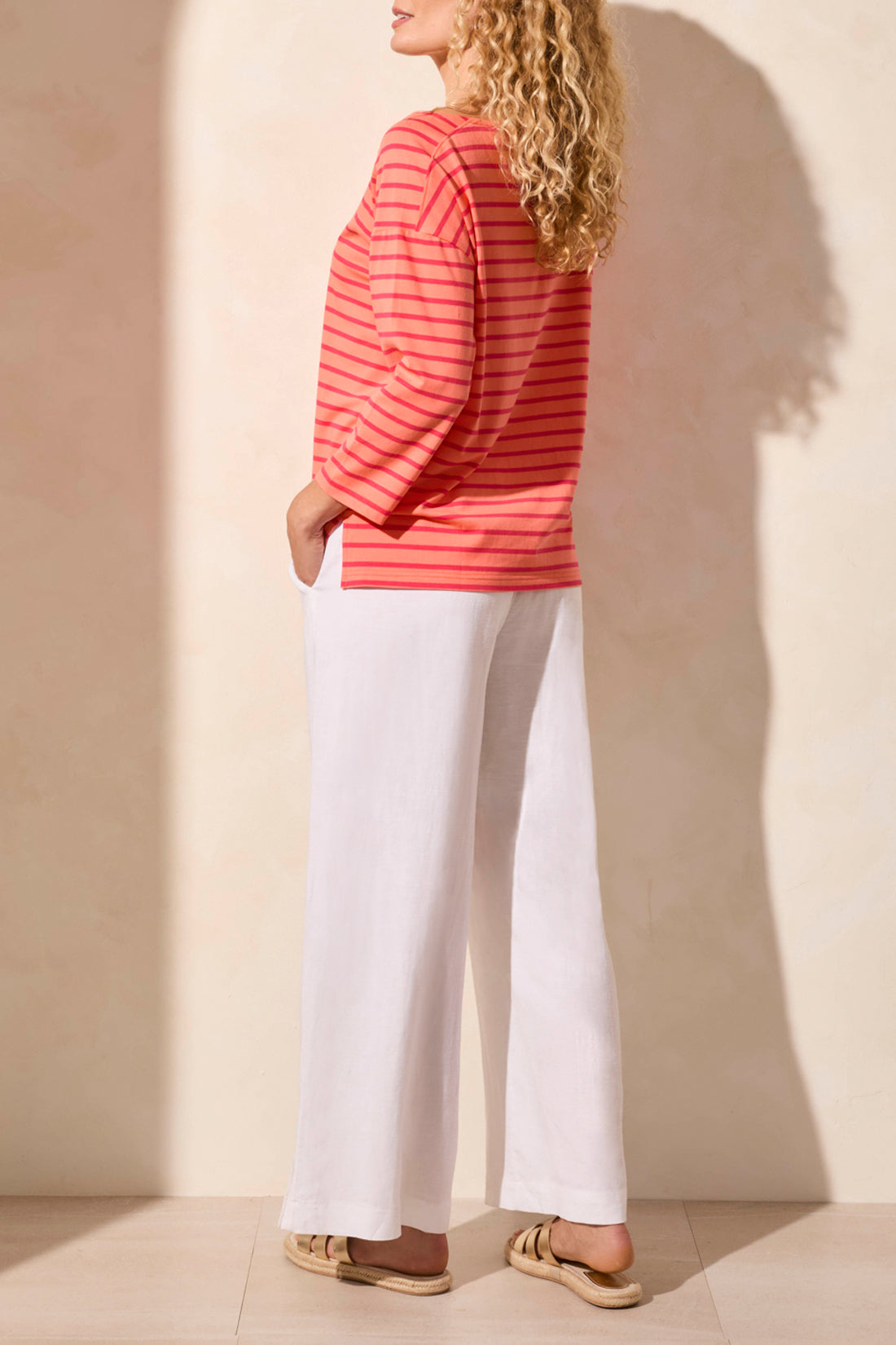 A person wearing a stylish Tribal 3/4 Sleeve Boat Neck Top in coral and pink stripes paired with white pants stands against a beige background.