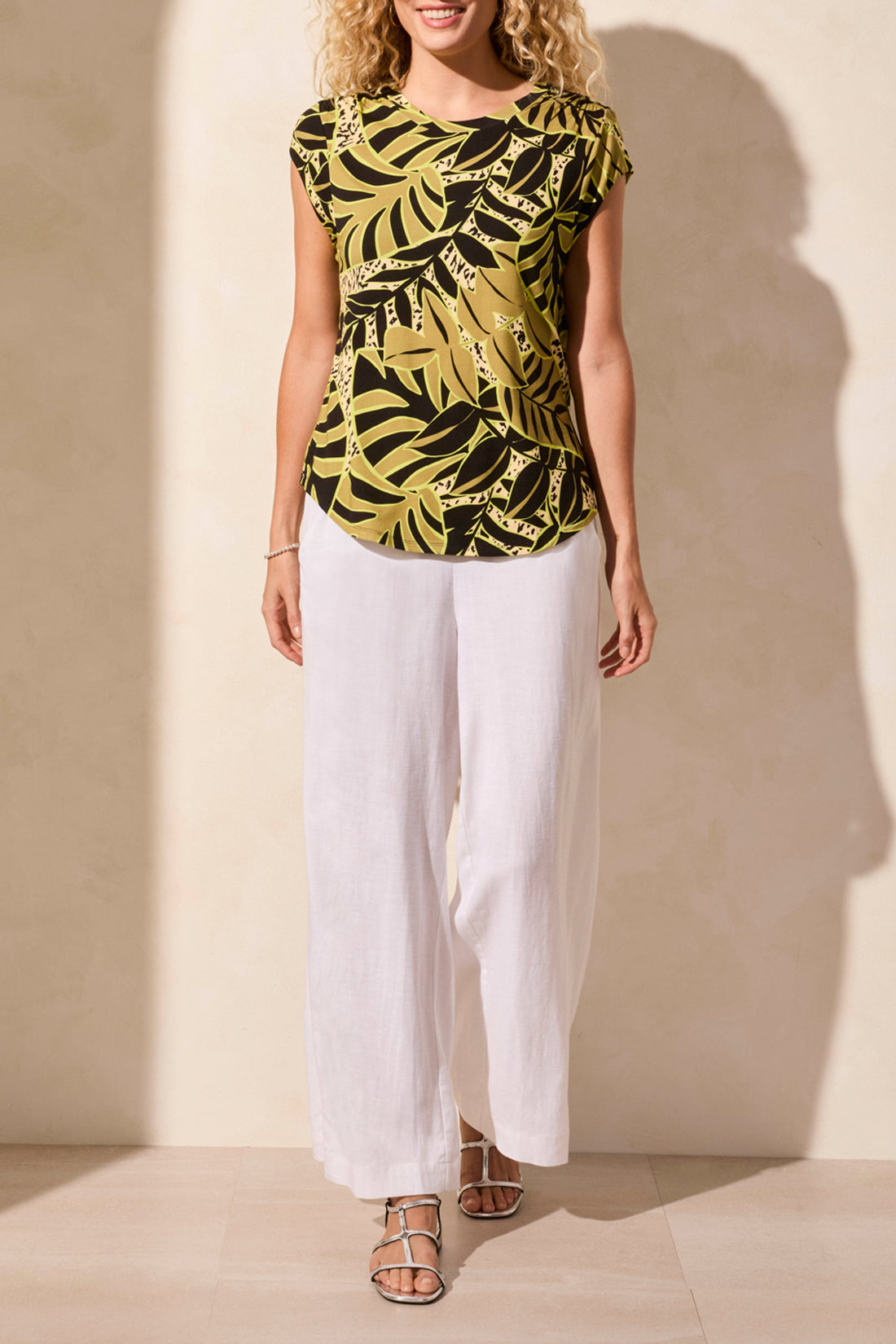 Against a light-colored background, a person wears Tribal's CAP SLEEVE TOP W/SHIRRING featuring a green and black tropical leaf print, paired with crisp white pants.