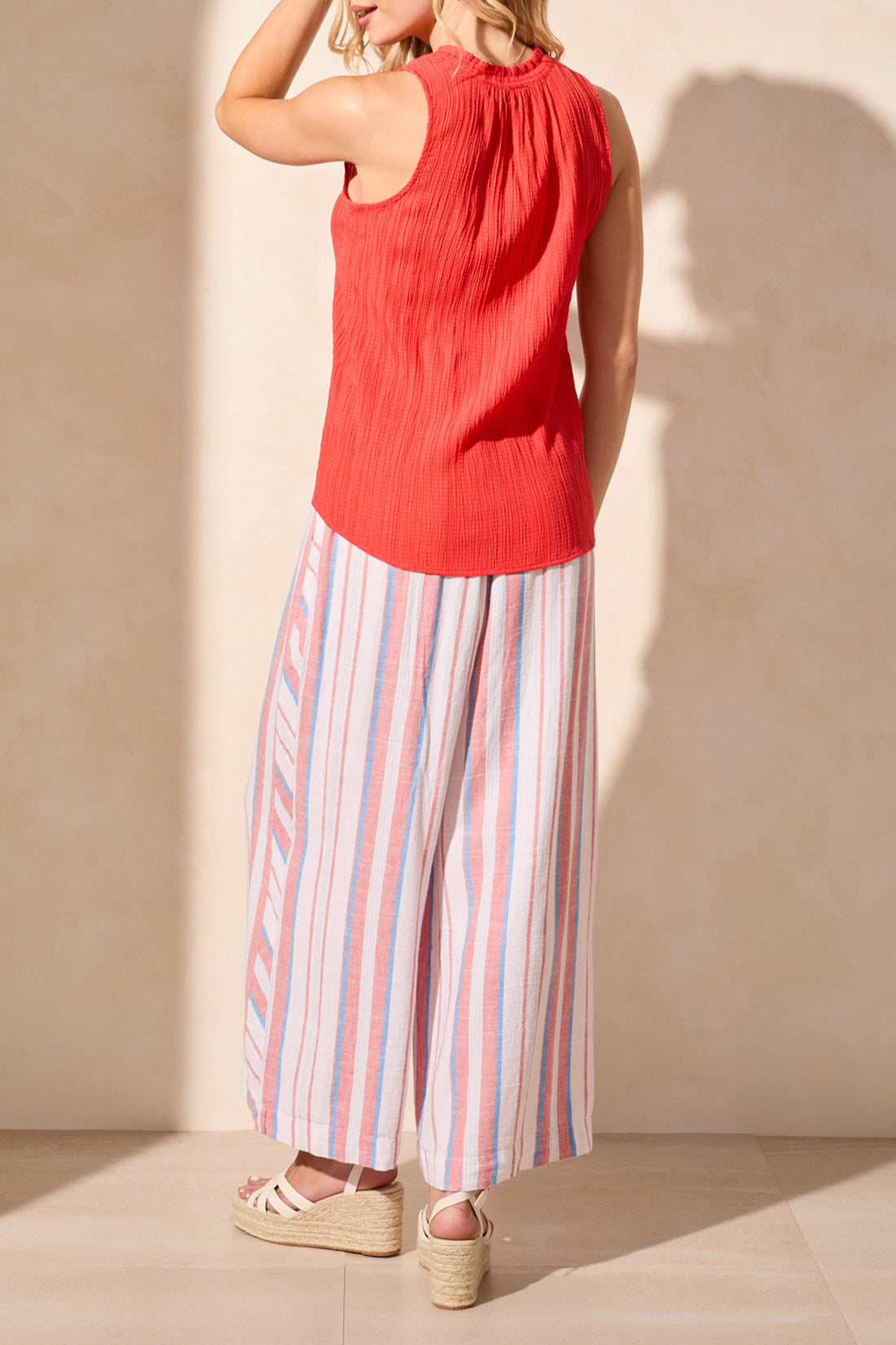 A woman models the Tribal brand's Sleeveless Blouse with Button Placket in red, paired with striped pants, standing against a neutral backdrop and embodying timeless style and elegance.